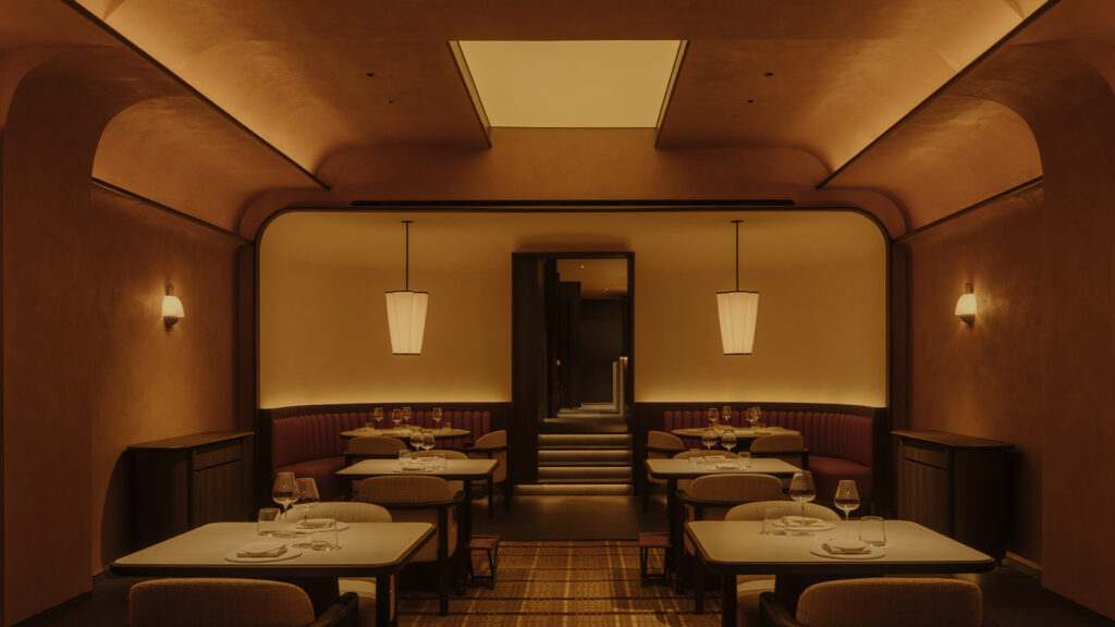 Warm, elegant restaurant dining room with softly lit tables, curved architectural details, and intimate amber lighting.